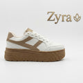 A low-top platform sneaker in white and tan, shown from the side against a light gray background. The brand name 'Zyra' with a leaf logo is displayed in the upper right corner.
